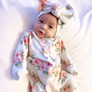 May include: A baby wearing a white floral print onesie with a ruffled neckline and a matching floral print headband. The onesie has long sleeves and a zipper closure.