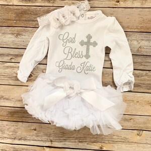 May include: White baby outfit with long sleeves and a silver glitter cross and the text "God Bless Giada Katie". The outfit is paired with a white tulle tutu with a white satin bow and a white flower headband.