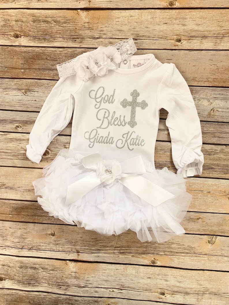 May include: White baby outfit with long sleeves and a silver glitter cross and the text "God Bless Giada Katie". The outfit is paired with a white tulle tutu with a white satin bow and a white flower headband.