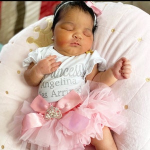 May include: A baby girl wearing a white onesie with the text "Princess Angelina Has Arrived" in silver glitter. She is wearing a pink tulle tutu with a large pink bow and a rhinestone crown.