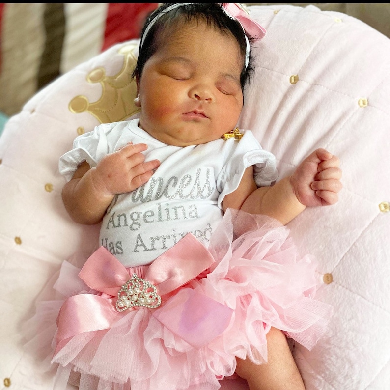 May include: A baby girl wearing a white onesie with the text "Princess Angelina Has Arrived" in silver glitter. She is wearing a pink tulle tutu with a large pink bow and a rhinestone crown.