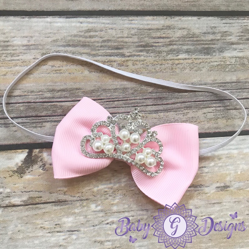 May include: A pink bow headband with a silver crown adorned with white pearls. The headband is made of elastic and is suitable for babies and toddlers.
