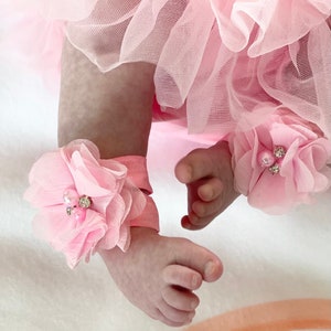 May include: Pink baby leg warmers with a flower design and pearl accents. The leg warmers are made of soft fabric and are perfect for keeping baby's feet warm and cozy.