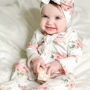May include: A baby wearing a white floral print onesie with a white bow headband. The onesie has a ruffled neckline and long sleeves. The baby is holding a wooden block with a footprint design.