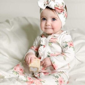 May include: A baby wearing a white floral print onesie with a white headband with a floral print bow. The baby is holding a wooden block with the text "08.2021" on it.