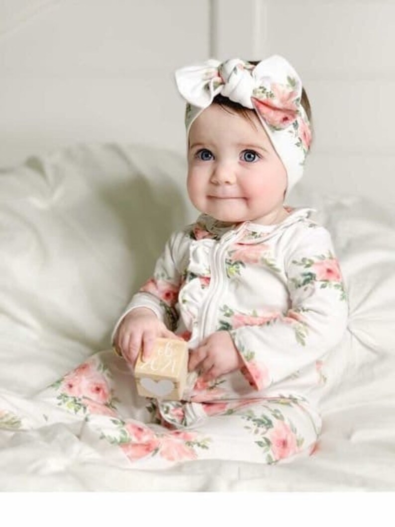 May include: A baby wearing a white floral print onesie with a white headband with a floral print bow. The baby is holding a wooden block with the text "08.2021" on it.