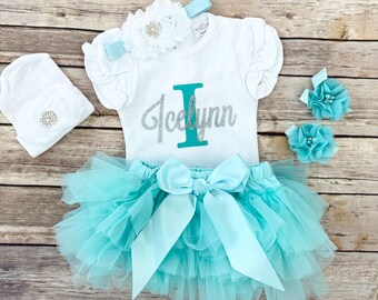 May include: A white baby outfit set with a tutu skirt, a shirt with the name "Icelynn", a headband, a beanie, and two hair clips. The tutu skirt and accessories are turquoise. The shirt has a turquoise "I" and the name "Icelynn" in silver glitter.