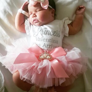 May include: A baby girl wearing a white onesie with the text "Princess Valentina Has Arrived" in silver glitter. She is wearing a pink tulle tutu with a pink satin bow and a crown-shaped brooch. The baby is wearing a pink headband with a crown and pearls.