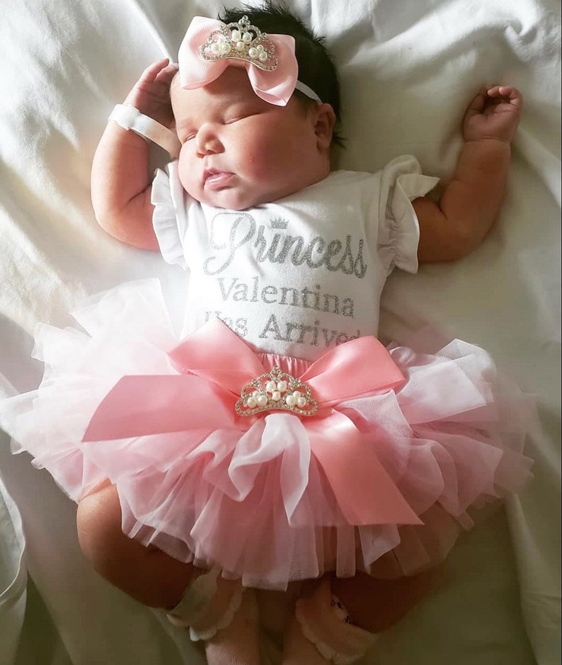 May include: A baby girl wearing a white onesie with the text "Princess Valentina Has Arrived" in silver glitter. She is wearing a pink tulle tutu with a pink satin bow and a crown-shaped brooch. The baby is wearing a pink headband with a crown and pearls.
