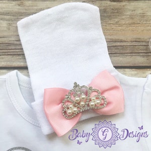 May include: A white baby hat with a pink bow and a silver crown with pearl accents. The bow has a rhinestone design.