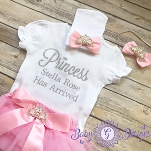 May include: A pink and white baby girl outfit with a white onesie that says "Princess Stella Rose Has Arrived" in silver glitter. The outfit also includes a pink tutu skirt, a pink headband with a pearl crown, and a pink bow with a pearl crown.