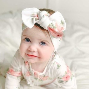May include: A baby wearing a white and pink floral print onesie with a white knotted headband. The headband has a large bow and a floral print.