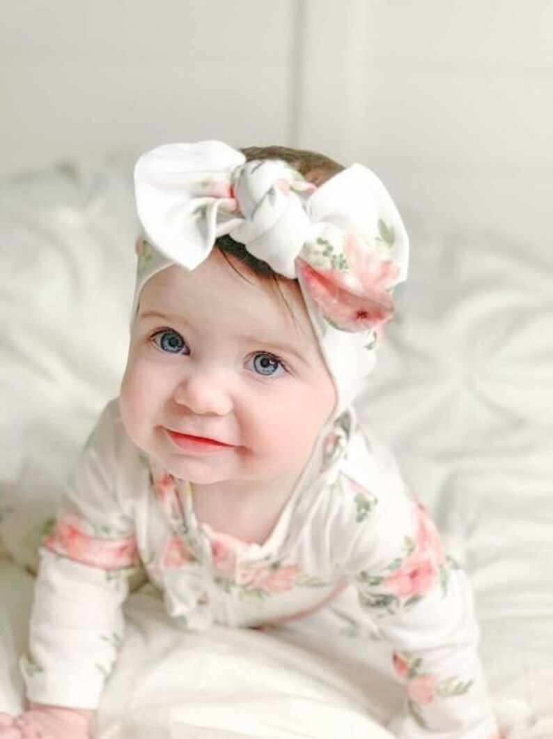 May include: A baby wearing a white and pink floral print onesie with a white knotted headband. The headband has a large bow and a floral print.