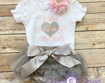 May include: A white baby outfit set with a tutu skirt, bloomers, and a headband. The shirt has the text "I Was Worth The Wait" with a glitter heart design. The tutu and bloomers are gray, and the bow is silver. A pink flower adorns the headband.