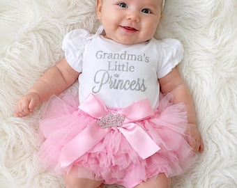 May include: A white short-sleeved bodysuit with the silver glitter text Grandma's Little Princess, paired with a pink ruffled tulle bloomer featuring a large satin bow and a silver crown embellishment, plus a matching pink floral headband.