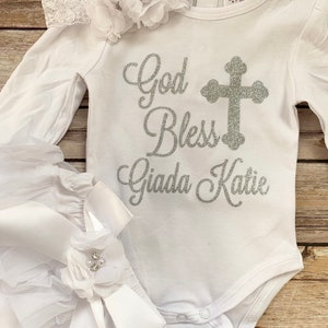 May include: White baby outfit with long sleeves and a white tulle skirt. The bodysuit has a silver glitter design that says "God Bless Giada Katie" with a silver glitter cross.