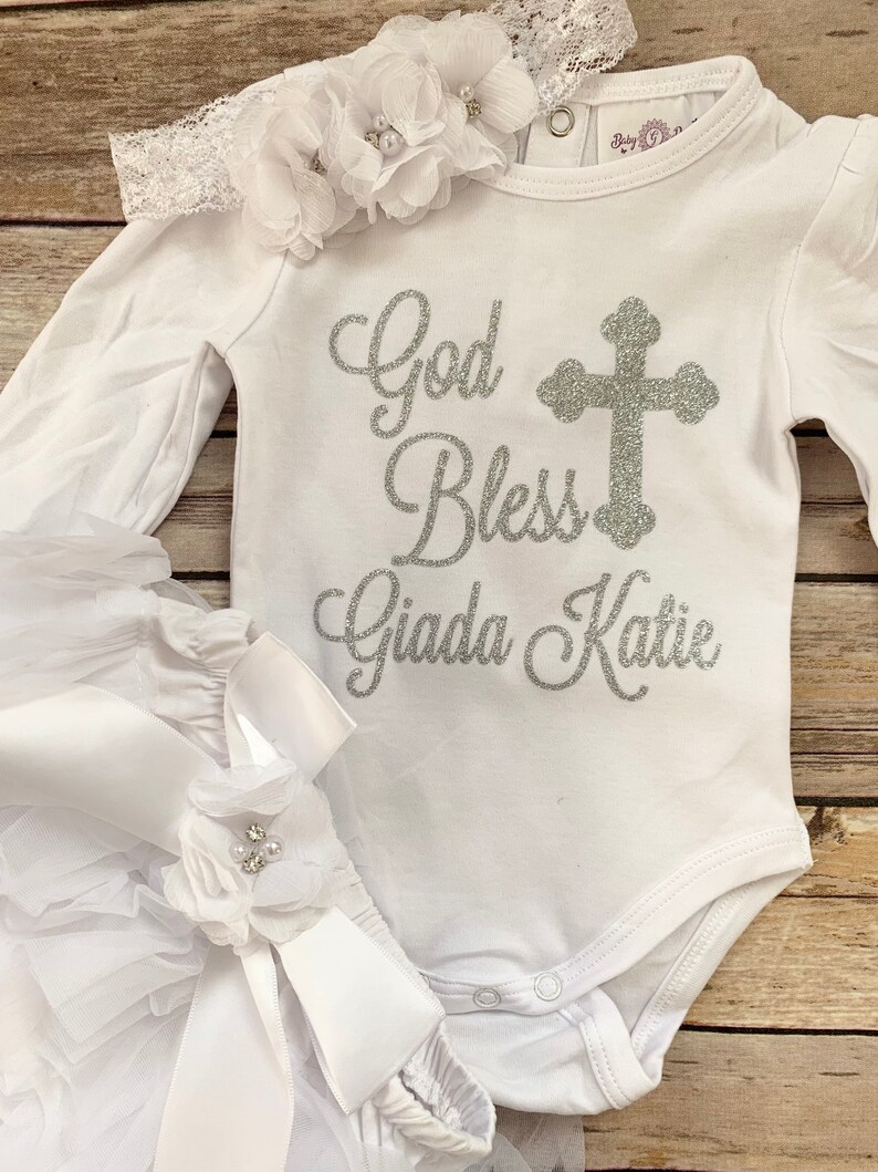 May include: White baby outfit with long sleeves and a white tulle skirt. The bodysuit has a silver glitter design that says "God Bless Giada Katie" with a silver glitter cross.