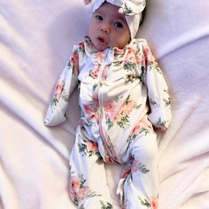 May include: A baby wearing a white floral print sleeper with a pink bow headband. The sleeper has a zipper closure and ruffled trim.