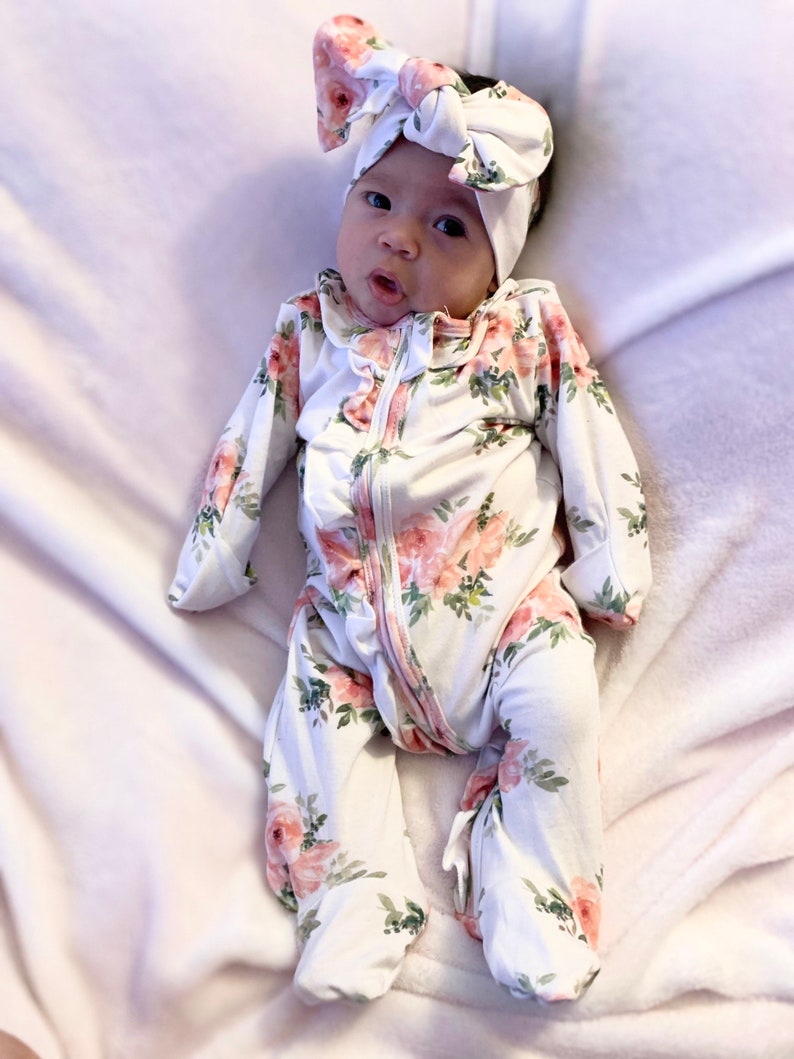 May include: A baby wearing a white floral print sleeper with a pink bow headband. The sleeper has a zipper closure and ruffled trim.