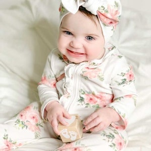 May include: A baby wearing a white and pink floral print onesie with a white bow headband. The baby is holding a wooden block with a footprint design.