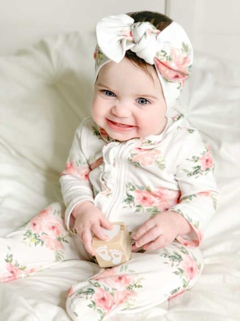 May include: A baby wearing a white and pink floral print onesie with a white bow headband. The baby is holding a wooden block with a footprint design.