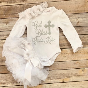 May include: White baby outfit with long sleeves, a white tulle skirt, and a headband with flowers. The shirt has a silver glitter cross and the text "God Bless Giada Katie".