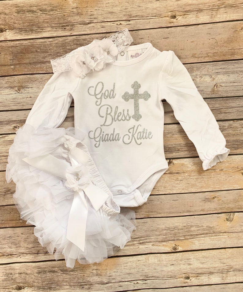 May include: White baby outfit with long sleeves, a white tulle skirt, and a headband with flowers. The shirt has a silver glitter cross and the text "God Bless Giada Katie".