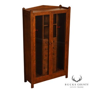 May include: A tall, brown wooden cabinet with two glass-paneled doors. The interior features vertical storage compartments, small drawers with round knobs, and red-accented supports. The top edge has a pointed, angular design with finials.