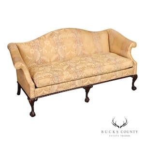 May include: A camelback sofa upholstered in a light gold fabric with a subtle floral and striped damask pattern. The piece features rolled arms, a single bench seat cushion, and dark wood legs carved with ball and claw feet details.