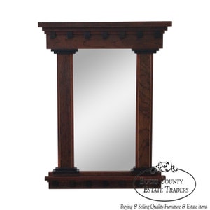 May include: A dark wood framed mirror with a simple design. The frame has two columns on either side of the mirror and a decorative top piece. The mirror is rectangular and has a beveled edge.