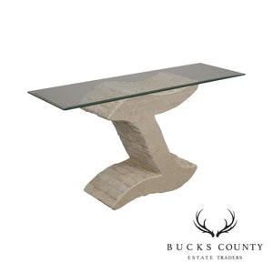 May include: A contemporary console table with a unique, abstract stone base and a clear glass top. The base is made of a light beige stone with a textured finish. The glass top is rectangular and sits on top of the base.