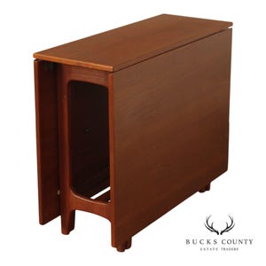 May include: A brown wooden drop-leaf table with a rectangular top and a smooth finish. The table features a central support structure with an open rectangular cutout and four legs. The side leaves are folded down vertically along the sides.