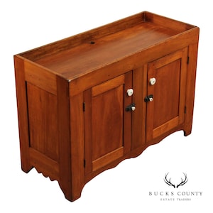 May include: A rectangular wooden washstand featuring a warm brown finish, a raised gallery edge on the top surface, and two cabinet doors. The doors are equipped with white ceramic knobs and black metal latches, with a scalloped base design.