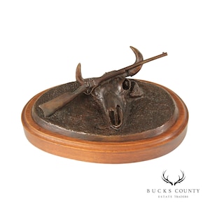 May include: A bronze sculpture of a bull skull with a rifle resting on top. The sculpture is mounted on a wooden base.
