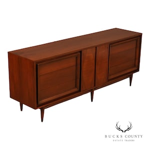 May include: A long, rectangular wooden sideboard featuring a rich brown finish. The piece includes two large cabinet sections with horizontal paneling and a central vertical panel, all supported by four tapered legs extending from the base.