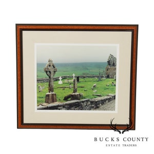 May include: A framed photograph of a cemetery in Ireland. The photo shows a stone Celtic cross, a ruined church, and a grassy field. The sky is overcast and the photo is in a brown wood frame.