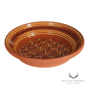 May include: A terracotta-colored ceramic bowl with a wide rim. The interior features a repeating pattern of black and yellow wavy lines and a central design of interlocking shapes. The bowl has a simple, functional design.