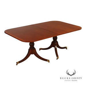 May include: A rectangular wooden dining table with a smooth, reddish-brown finish. The table features two central pedestal bases, each with four curved legs ending in brass-toned metal casters. The surface shows a clear seam where two sections meet.