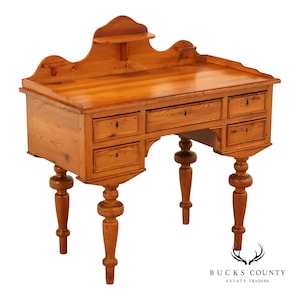 May include: A light brown wooden desk featuring a raised back gallery with two small shelves, a central drawer, and four smaller side drawers. The desk is supported by four turned legs with bulbous detailing and includes keyholes on each drawer front.