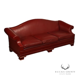 May include: A deep red leather sofa featuring a curved backrest, rolled arms, and three removable seat cushions. The base is supported by four wooden feet, and the frame is accented with decorative nailhead trim along the lower front edge.