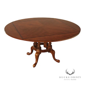 May include: A round wooden dining table with a rich brown finish and a square inlay pattern on the tabletop surface. The table is supported by a central pedestal base featuring four curved legs with carved details and a circular support ring.