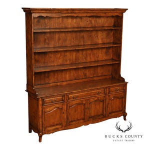 May include: A tall wooden hutch with a warm brown finish, featuring three open upper shelves and a base cabinet with three drawers and three lower doors. The piece has a decorative arched top molding and curved feet, with visible wood grain.