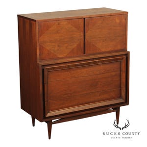 May include: A tall wooden chest of drawers featuring a warm brown finish. The upper section has two cabinet doors with a diamond-shaped wood grain pattern, while the lower section contains three horizontal drawers. The piece stands on four tapered legs.