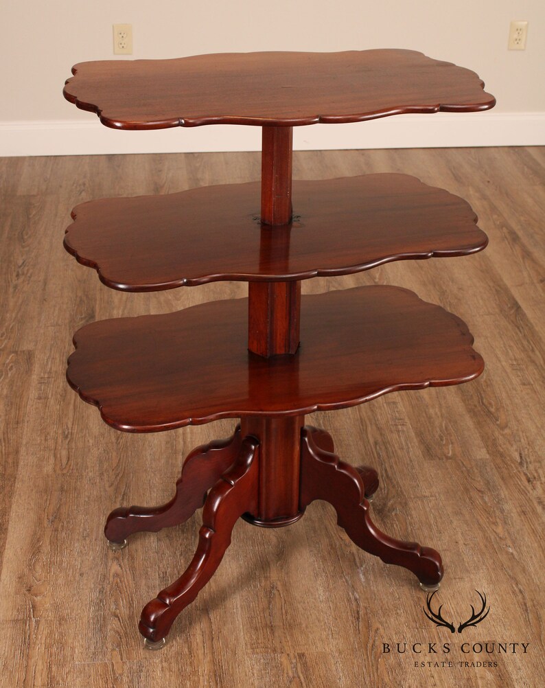 May include: A three-tiered mahogany table with a scalloped edge on each shelf. The table has a turned pedestal base with four carved legs.