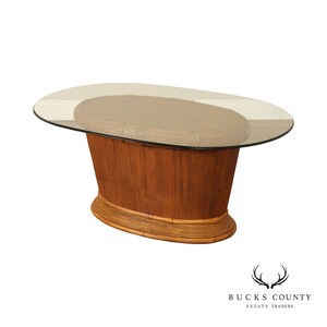 May include: Oval-shaped, glass-top coffee table with a woven rattan base. The base is a light brown color and has a rounded, oval-shaped design.