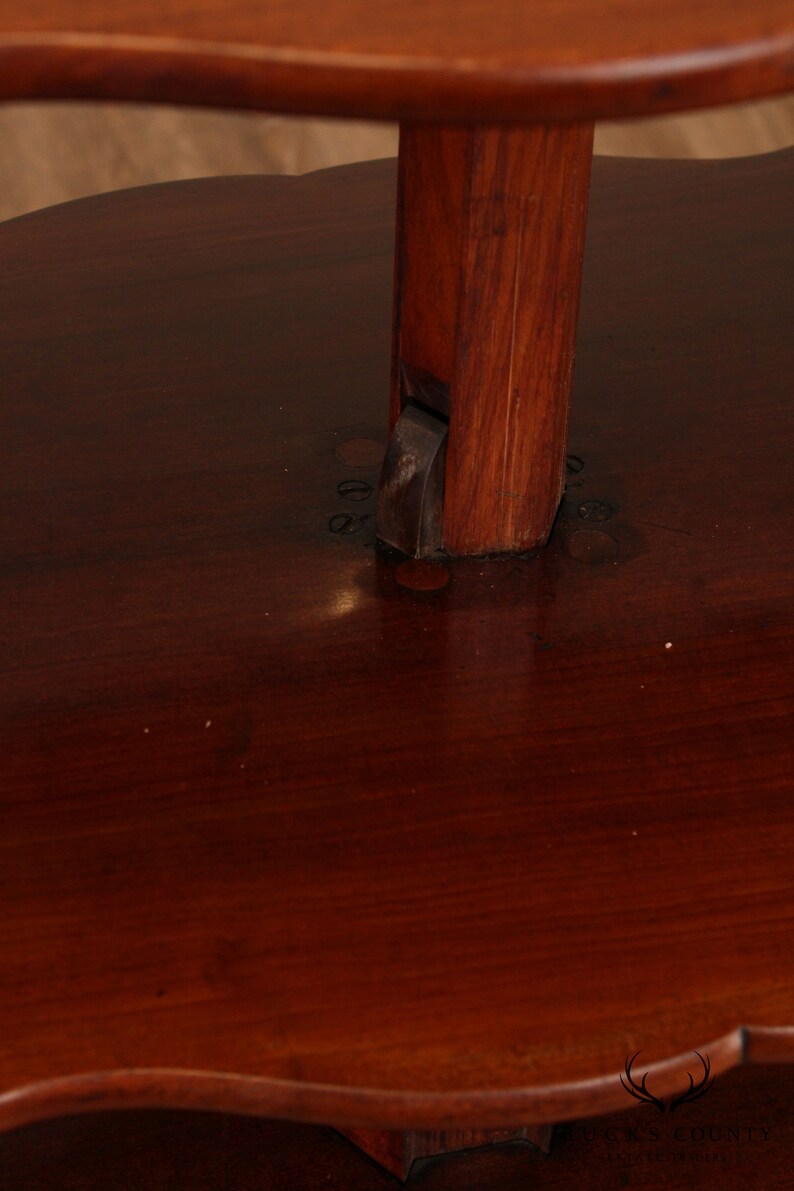 May include: A two-tiered wooden table with a dark brown finish. The table has a round top and a scalloped edge. The bottom tier is slightly smaller than the top tier. The table is supported by a single, sturdy wooden post.