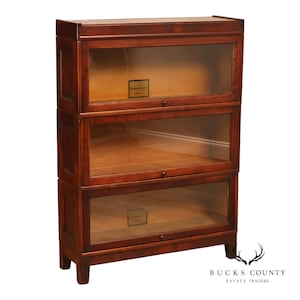 May include: A three-tier mahogany barrister bookcase featuring a rich reddish-brown wood finish. Each of the three sections includes a clear glass drop-front door with a small wooden knob. A rectangular label is visible on the glass of the top shelf.