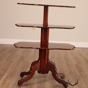 May include: A three-tiered mahogany wood table with a scalloped edge on each shelf. The table has a turned pedestal base with four cabriole legs and brass casters.