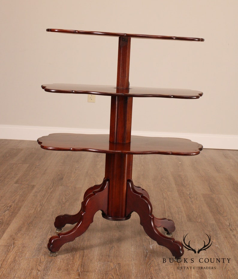 May include: A three-tiered mahogany wood table with a scalloped edge on each shelf. The table has a turned pedestal base with four cabriole legs and brass casters.