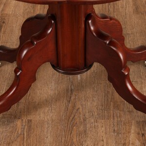 May include: Close-up of the base of a mahogany table with three legs. Each leg has a carved design and ends in a rounded foot with a brass caster.